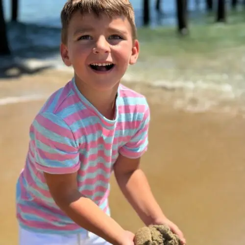 HB1452b-2 – Togs Moments Smiling boy wearing a pink and gray striped tee at the beach, available at Togs – GCC kidswear reseller for casual summer fashion +Post Title + Separator + Category + Separator + Site Title