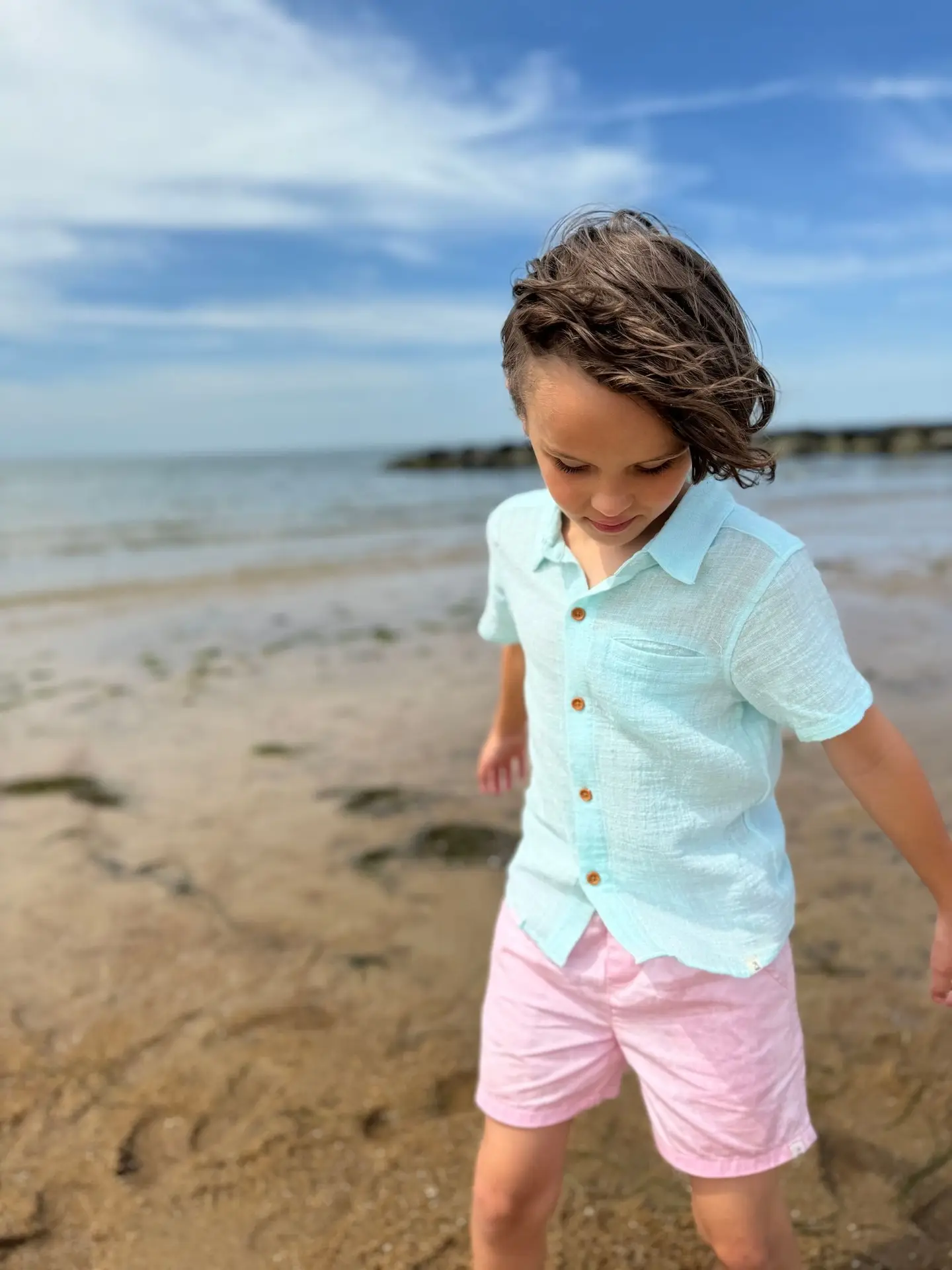 HB1474r-2 – Togs Moments Boy wearing a light blue button-up shirt and pink shorts walking on the beach, outfit available at Togs – GCC kidswear reseller for coastal summer fashion +Post Title + Separator + Category + Separator + Site Title