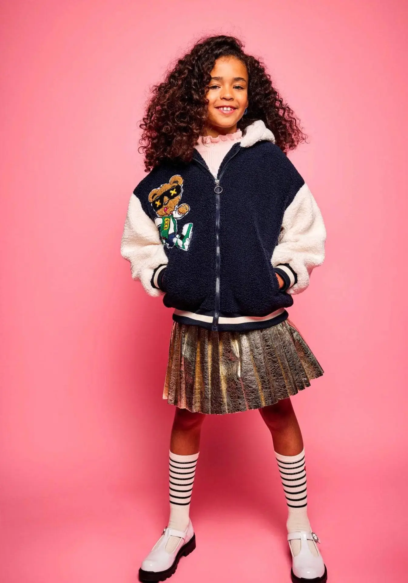 FoilPleatedSkirt-9 – Togs Moments Confident girl wearing a foil pleated skirt and varsity jacket with bear patch, available at Togs – GCC reseller for stylish girls fashion +Post Title + Separator + Category + Separator + Site Title